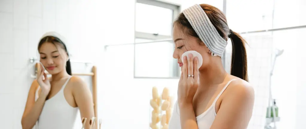 What Is the Best Age to Get Botox? Woman in a white tank top, who recently joined a skincare-focused healthcare membership, applies product with a cotton pad in front of the bathroom mirror, wearing a headband.