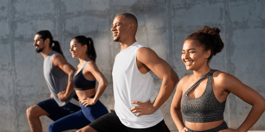 How Frequently Should You Work With a Personal Trainer? Four people in athletic wear exercise outdoors, standing in a row and smiling while performing lunges against a concrete wall—showcasing the confidence that comes from healthy living and Medical-Grade Skin Care.